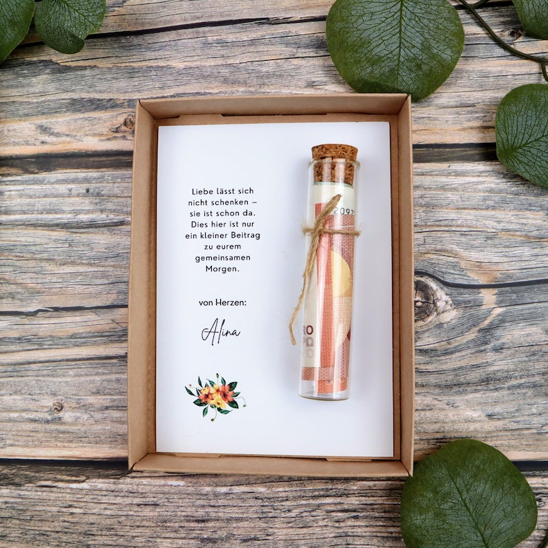 May include: A brown cardboard gift box containing a white card with German text and a small floral illustration. A clear glass vial with a cork stopper is tied to the card with twine, containing a rolled-up banknote inside the glass container.