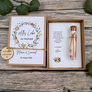 May include: A brown cardboard gift box containing a wedding card with a floral wreath, the text Alles Liebe zur Hochzeit, Maria & Leonard, and 22, August 2026. A small wooden tag and a glass vial with a cork stopper containing a rolled paper note.
