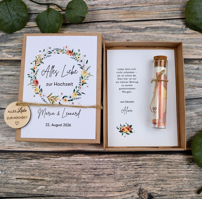 May include: A brown cardboard gift box containing a wedding card with a floral wreath, the text Alles Liebe zur Hochzeit, Maria & Leonard, and 22, August 2026. A small wooden tag and a glass vial with a cork stopper containing a rolled paper note.