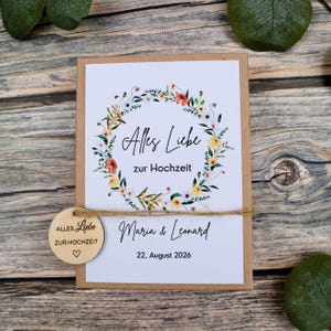 May include: A rectangular card with a white background featuring a watercolor floral wreath, the text Alles Liebe zur Hochzeit, the names Maria & Leonard, and the date 22, August 2026. A round wooden tag with the same text is tied with twine.