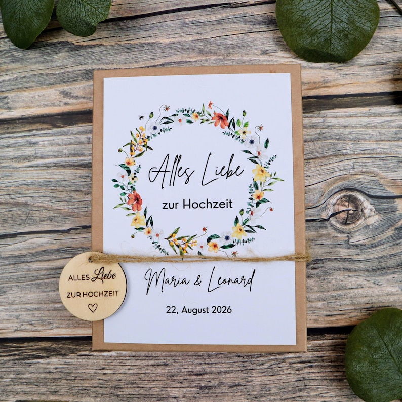 May include: A rectangular card with a white background featuring a watercolor floral wreath, the text Alles Liebe zur Hochzeit, the names Maria & Leonard, and the date 22, August 2026. A round wooden tag with the same text is tied with twine.