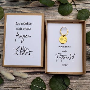 Puede incluir: Dos cajas de regalo con texto en alem&aacute;n. Una caja dice "Ich m&ouml;chte dich etwas fragen" con una ilustraci&oacute;n de choque de pu&ntilde;os. La otra caja contiene un llavero dorado que dice "Patenonkel" y pregunta "M&ouml;chtest du mein Patenonkel sein?"