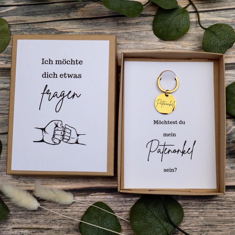 Puede incluir: Dos cajas de regalo con texto en alem&aacute;n. Una caja dice "Ich m&ouml;chte dich etwas fragen" con una ilustraci&oacute;n de choque de pu&ntilde;os. La otra caja contiene un llavero dorado que dice "Patenonkel" y pregunta "M&ouml;chtest du mein Patenonkel sein?"