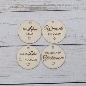 May include: Four circular light-colored wooden tags, each featuring a small hole at the top and a laser-engraved heart. The tags display the German phrases: EIN LEBEN LANG, WUNSCH ERF&Uuml;LLER, ALLES LIEBE ZUR HOCHZEIT, and HERZLICHEN GL&Uuml;CKWUNSCH.