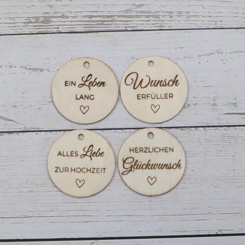 May include: Four circular light-colored wooden tags, each featuring a small hole at the top and a laser-engraved heart. The tags display the German phrases: EIN LEBEN LANG, WUNSCH ERF&Uuml;LLER, ALLES LIEBE ZUR HOCHZEIT, and HERZLICHEN GL&Uuml;CKWUNSCH.