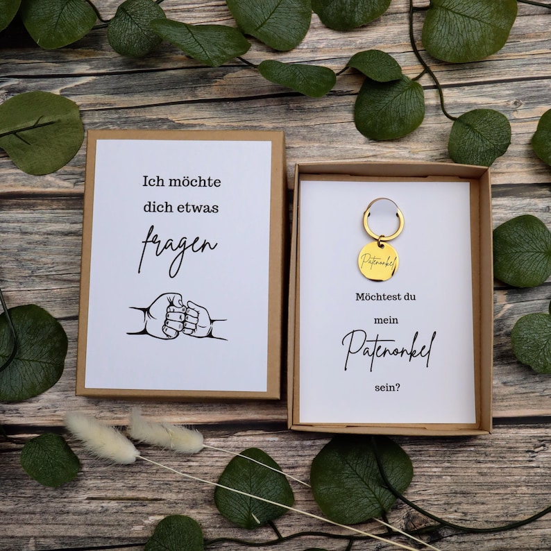 Puede incluir: Un juego de regalo con una tarjeta y un llavero. La tarjeta dice "Ich m&ouml;chte dich etwas fragen" con un gr&aacute;fico de manos entrelazadas. El llavero es dorado y tiene la palabra "Patenonkel". La segunda tarjeta dice "M&ouml;chtest du mein Patenonkel sein?"