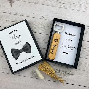 May include: A black box with a white card inside that says "Hol die Flege raus!" and "Du wirst gebraucht!" with a black bow tie illustration. A wooden bottle opener with the text "Trauzeuge" is in a black box with a white card that says "Willst du mein Trauzeuge sein?"