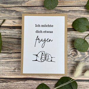 Puede incluir: Una tarjeta de felicitaci&oacute;n con fondo blanco y borde marr&oacute;n. La tarjeta tiene el texto alem&aacute;n "Ich m&ouml;chte dich etwas fragen" y un gr&aacute;fico de dos pu&ntilde;os chocando. La tarjeta est&aacute; sobre una superficie de madera con hojas verdes.