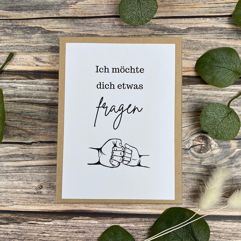 Puede incluir: Una tarjeta de felicitaci&oacute;n con fondo blanco y borde marr&oacute;n. La tarjeta tiene el texto alem&aacute;n "Ich m&ouml;chte dich etwas fragen" y un gr&aacute;fico de dos pu&ntilde;os chocando. La tarjeta est&aacute; sobre una superficie de madera con hojas verdes.