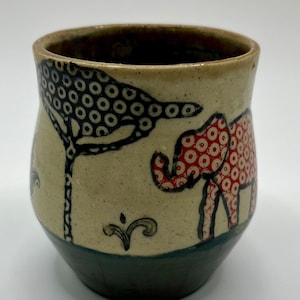 Puede incluir: Una taza de cerámica con un diseño pintado a mano de un elefante y un árbol. El elefante es rojo con lunares blancos y el árbol es negro con lunares blancos. La taza tiene un esmalte azul oscuro en la parte inferior.