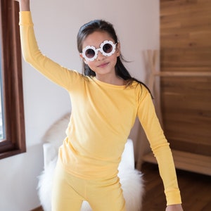 May include: A young person wearing a yellow long-sleeved shirt and matching pants. They are wearing white flower-shaped sunglasses and have long dark hair.