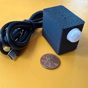May include: A small, black electronic device with a white sensor and a black USB cable. A penny is next to the device for size comparison. The device is on a yellow surface.