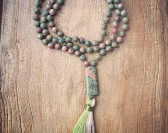 May include: A beaded necklace with green and red-toned beads. The necklace has a rectangular stone pendant and a tassel with green, white, and pink threads. The necklace is displayed on a wooden surface.