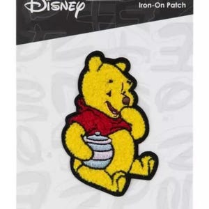May include: An iron-on patch featuring Winnie the Pooh, a yellow bear wearing a red shirt, holding a blue and white honey pot. The patch is on a white background with the Disney logo and the words "Iron-On Patch".