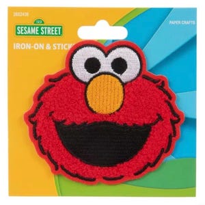 May include: A red, iron-on and stick-on patch featuring the Sesame Street character Elmo. The patch has a textured, plush appearance with embroidered details. The background is yellow and blue, with the words "Sesame Street" and "Paper Crafts" visible.