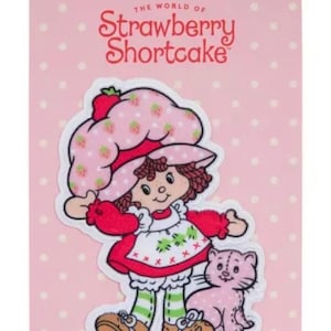 May include: A Strawberry Shortcake patch featuring the character with a pink cat. The patch has a pink background with white polka dots. The character wears a pink hat with strawberries, a red dress, and green striped socks. The text "The World of Strawberry Shortcake" is at the top.