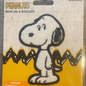 May include: A white and black Snoopy iron-on patch and sticker on a rectangular cardboard backing. The packaging features the text PEANUTS, IRON-ON & STICKER, and PAPER CRAFTS. The background has a yellow zig-zag pattern and a comic strip print.
