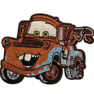 May include: An embroidered iron-on patch featuring a rusty brown tow truck character with a wide, toothy grin, large white eyes, and light blue paint accents. The truck has black tires, a prominent front bumper, and a rear-mounted towing crane.