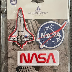May include: Three iron-on patches featuring NASA logos. The patches are white with red and blue embroidery. The first patch is a space shuttle, the second is a circle with a red orb and the text "NASA" in white, and the third is a rectangular patch with the text "NASA" in red.
