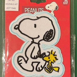 May include: An iron-on patch featuring Snoopy and Woodstock from the Peanuts comic strip. The patch has a white background with black outlines and a light blue border. Woodstock is yellow. The patch is on a red backing card.