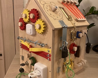 May include: A wooden toy house with a variety of activities for children, including a clock, gears, a zipper, a lock, a chain, a light switch, and an electrical outlet. The house is painted in natural wood tones.