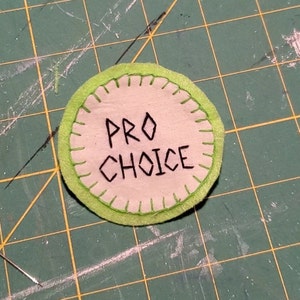 May include: A round, green-trimmed fabric patch with the words "PRO CHOICE" embroidered in black thread.