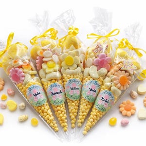Puede incluir: Cinco bolsas de cono transparentes llenas de caramelos amarillos y dulces variados, atadas con cintas amarillas. Cada bolsa tiene una etiqueta que dice "Felices Pascuas" y un nombre. Caramelos en forma de flor y corazón esparcidos.