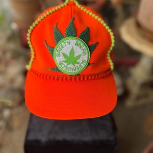 May include: Bright orange baseball cap with a green cannabis leaf patch and the words "SAFETY BREAK SUPERVISOR" in a circular design. The cap has a yellow trim.