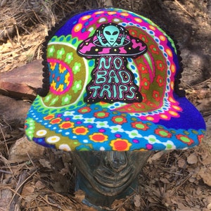 May include: A colorful, patterned hat with a black trim and the text "NO BAD TRIPS". The hat features a psychedelic design with an alien in a UFO. The hat is a vibrant mix of blue, green, pink, orange, and yellow.