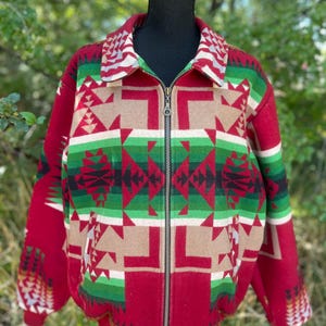 May include: A zip-up jacket featuring a bold geometric pattern in shades of red, green, tan, black, and white. The garment has a fold-down collar, a full-length metal zipper closure, and ribbed cuffs and hem, showcasing a symmetrical design.