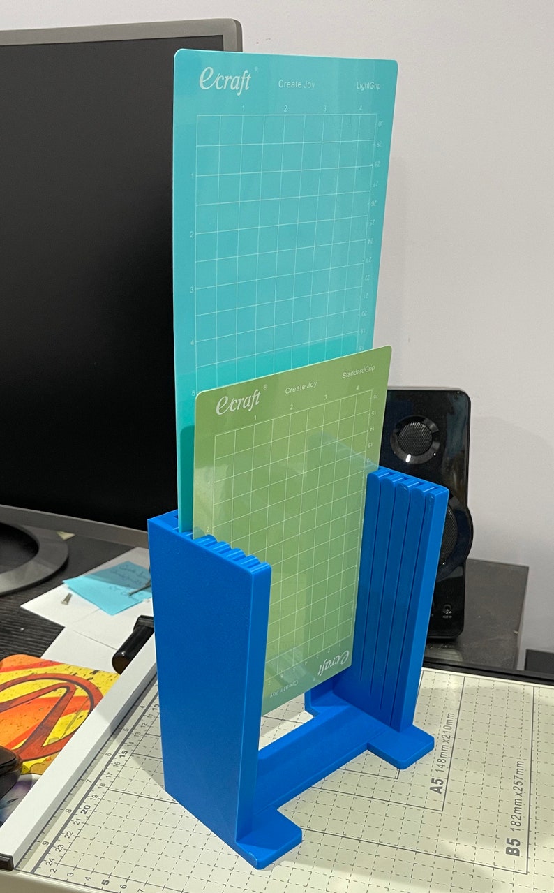 May include: A blue plastic stand holds two cutting mats. The top mat is light blue with the text "ecraft Create Joy LightGrip". The bottom mat is green with the text "ecraft Create Joy StandardGrip".