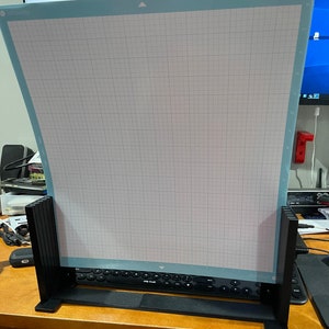 May include: A black stand with a light blue cutting mat with a grid pattern. The stand has a black base and black vertical supports. The cutting mat is designed for use with a craft cutting machine.
