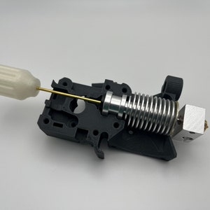 May include: A 3D printer hot end assembly, featuring a black plastic frame, a silver-colored heat sink, and a brass nozzle. A white plastic handle is connected to a brass rod. The components are set against a white background.