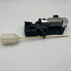 May include: A 3D printer hot end assembly with a black plastic housing, silver heat sink, and a brass nozzle. A white plastic cleaning tool with a brass rod is also included.
