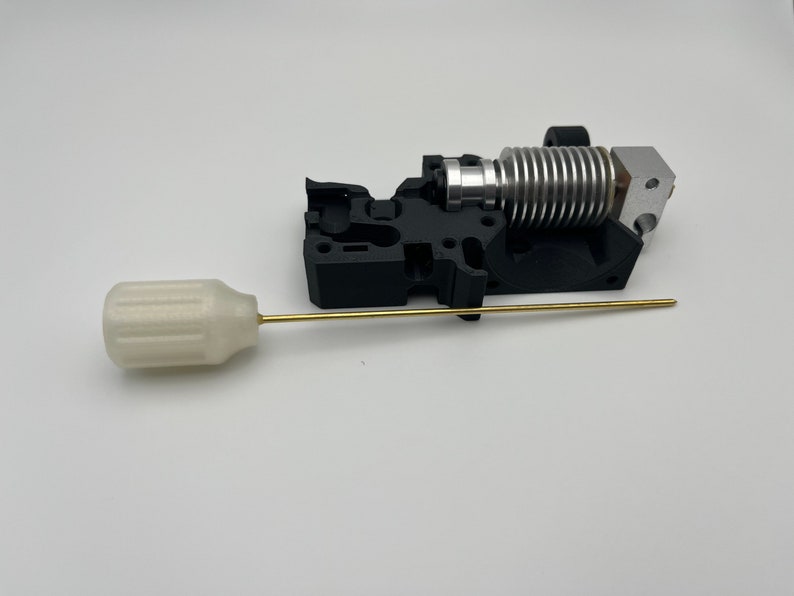 May include: A 3D printer hot end assembly with a black plastic housing, silver heat sink, and a brass nozzle. A white plastic cleaning tool with a brass rod is also included.
