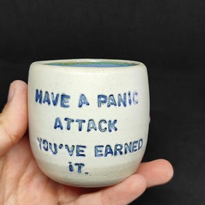Peut inclure: Une tasse en céramique blanche avec une inscription bleue qui dit "Have a panic attack you've earned it."