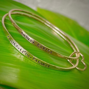 May include: Two gold bangle bracelets with engraved text. The top bracelet reads "Ku'ulei Kawika" and the bottom bracelet reads "Kauila Li'ua Pakamia".