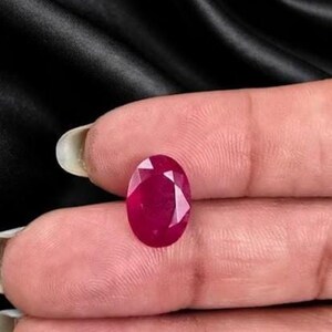 Puede incluir: Una gema de rubí de corte ovalado con un tono rojo intenso y una superficie multifacetada. La piedra muestra un acabado brillante y pulido con facetas geométricas claras que reflejan la luz, destacando su color intenso y su estructura cristalina transparente.