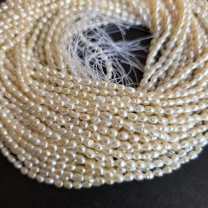 May include: A collection of creamy white, oval-shaped pearls strung together. The pearls are tightly arranged, forming a coiled pattern. The strands of pearls are on a dark background, with the stringing material visible at the centre.