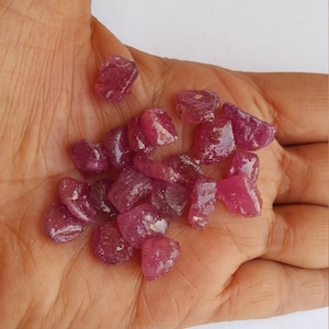 May include: A collection of small, rough, pink ruby gemstones. The rubies are various shapes and sizes, and have a translucent appearance.