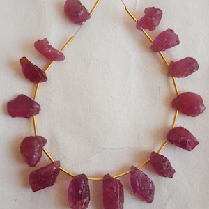 Op de afbeelding: Een streng roze robijn edelsteen kralen geregen aan een gouden draad. De kralen zijn ruw en onregelmatig van vorm, sommige hebben een puntige of gekartelde rand.