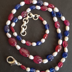 May include: A necklace featuring a combination of red, blue, and white beads. The necklace has a silver clasp and is composed of oval-shaped red and blue gemstones, interspersed with white pearls. The beads are strung together to form a necklace.