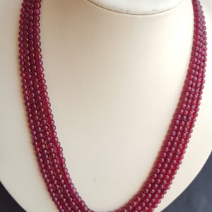 Op de afbeelding: Een ketting met meerdere strengen, samengesteld uit kleine, ronde, dieprode kralen. De ketting wordt getoond op een witte mannequin buste, waarbij de manier waarop de strengen hangen wordt benadrukt. De kralen hebben een gepolijste uitstraling.