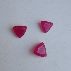 May include: Three triangular, faceted, ruby-red gemstones. Each gem has a small hole drilled near one point. The gems are displayed on a white surface, showcasing their vibrant colour and geometric shape. Ideal for jewellery making.