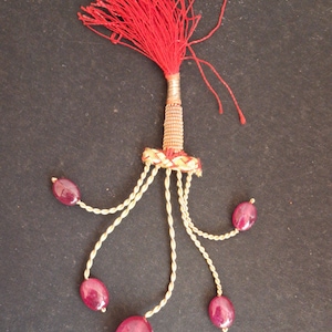 May include: A red tassel with a gold base and five strands of gold beads with pink gemstones hanging from the base.