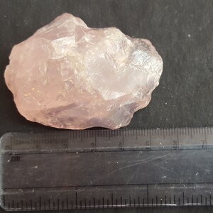 Peut inclure: Un gros cristal de quartz rose brut, translucide et de couleur rose p&acirc;le. Le cristal mesure environ 5 cm de long et est pos&eacute; sur une surface noire &agrave; c&ocirc;t&eacute; d'une r&egrave;gle.