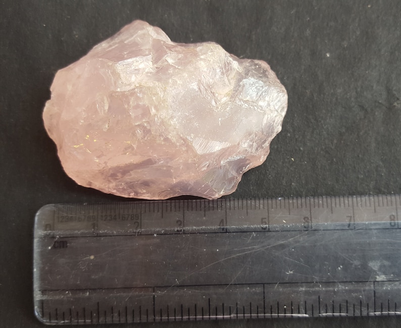 Peut inclure: Un gros cristal de quartz rose brut, translucide et de couleur rose p&acirc;le. Le cristal mesure environ 5 cm de long et est pos&eacute; sur une surface noire &agrave; c&ocirc;t&eacute; d'une r&egrave;gle.