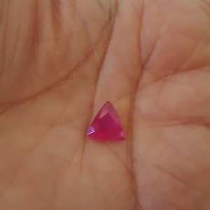 May include: A triangular, ruby-red gemstone with multiple facets. The gem is held in a hand, highlighting its vibrant colour and geometric shape. The facets reflect light, creating a sparkling effect.