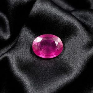 May include: A vibrant, oval-cut gemstone with a deep magenta-pink hue and multifaceted surface. The stone features a brilliant, reflective clarity with numerous geometric facets that catch the light, creating a sharp, symmetrical appearance.