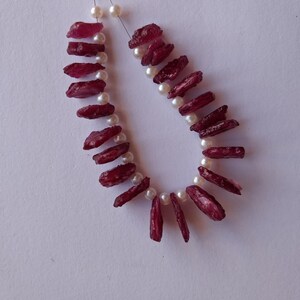 May include: A strand of red gemstone beads with white pearl spacers. The beads are irregular in shape and have a rough, natural texture.